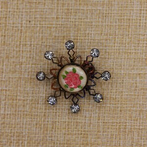 May include: A circular brooch featuring a central painted pink rose on a cream background, surrounded by a dark metal frame with radiating wire spokes. Each spoke is tipped with a small, clear, round faceted rhinestone accent.