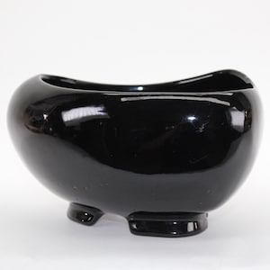 May include: A black ceramic planter with a rounded, oval shape and three short legs. The planter has a glossy finish.