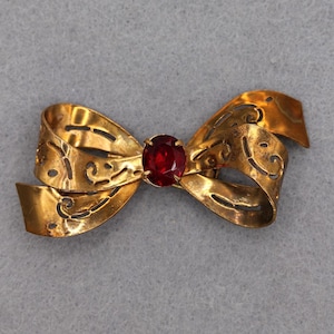 May include: A gold-toned metal brooch shaped like a bow with cut-out scrollwork patterns throughout the ribbon. The center of the bow features a single, round, faceted deep red gemstone held in place by four small metal prongs.