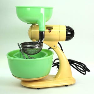 May include: Vintage Nutmaster food mill in cream and green. The appliance features a green hopper and bowl, a cream-colored motor housing, and a black motor end. A metal strainer and stirring tool are included. The cord is black.