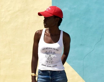 May include: A woman wearing a white tank top with the text "Let me hear you say Jesus, Jesus, Jesus." and a red baseball cap. She is standing in front of a yellow and blue wall.