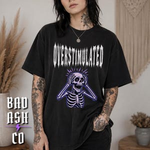 May include: A black short-sleeve t-shirt featuring the white text OVERSTIMULATED in a jagged font above a graphic of a purple and white skeleton with its hands on its head, appearing to be in distress, centred on the chest of the garment.