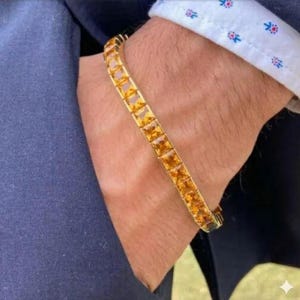 Könnte beinhalten: Ein goldenes Armband mit rechteckigen, bernsteinfarbenen Edelsteinen. Das Armband wird am Handgelenk getragen, mit einer marineblauen Jacke und einem weißen Hemd mit floralem Muster im Hintergrund.
