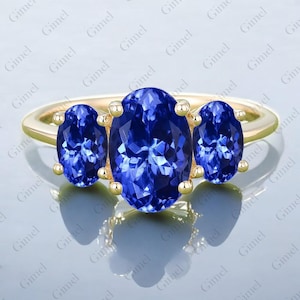 May include: A gold ring featuring three oval-cut, deep blue gemstones. The central stone is larger than the two flanking it. The ring's band is a simple, polished gold, and the gemstones are set with small gold prongs.