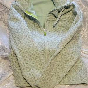 May include: A light green zip-up hoodie with a repeating starburst pattern. The hoodie has a hood, a full-length zipper, and a lime green accent on the zipper. The interior is a lighter shade of green. The hoodie is made of a soft material.