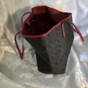 Puede incluir: Un bolso tote de color marrón oscuro con un patrón de logotipo circular repetitivo en el exterior. El bolso tiene un forro interior de color rojo vibrante y un borde rojo a lo largo de la abertura superior. Correas rojas finas están unidas a los lados.
