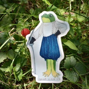 May include: A sticker featuring a green frog dressed in a white shirt, blue skirt, and black gloves, holding a sword. The frog has yellow feet and is standing in a grassy area. The sticker has the text "JRGES 6.20.23".