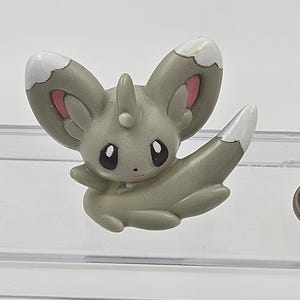 May include: A small, grey, plastic figurine of a Pokemon character with white and pink accents. The character has large ears, a pointed tail, and a small horn on its head.