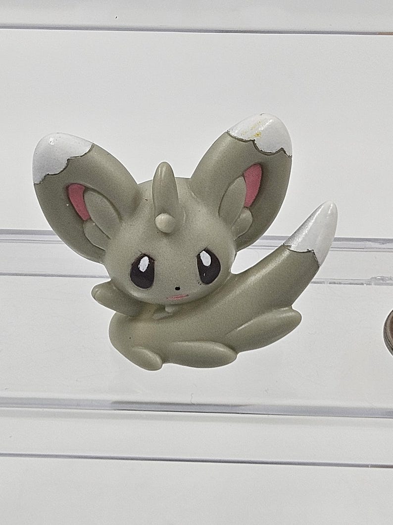 May include: A small, grey, plastic figurine of a Pokemon character with white and pink accents. The character has large ears, a pointed tail, and a small horn on its head.