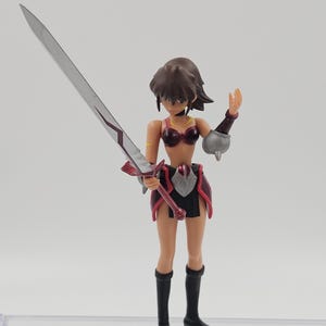 May include: A warrior figurine with a silver sword, clad in a burgundy and black outfit. The figure has brown hair and black boots. A silver coin is on the surface.