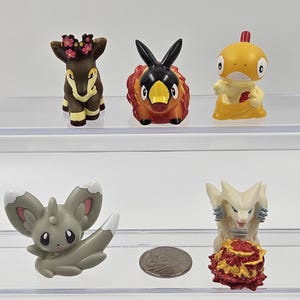 May include: Five small plastic figurines of Pokemon characters. The characters are a brown deer with a flower crown, a red and black fire-type Pokemon, a yellow and orange water-type Pokemon, a grey and white rabbit-like Pokemon, and a white and red dragon-like Pokemon.
