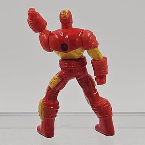May include: A small, molded plastic figurine of a character in a red and gold armored suit. The figure is shown from the back, with one arm raised in a fist, featuring a circular indentation on the upper back and textured gold knee pads.
