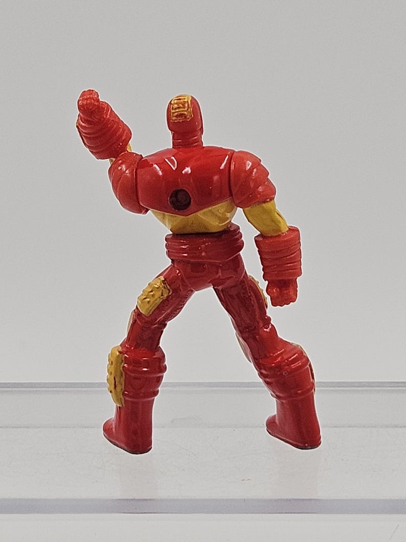 May include: A small, molded plastic figurine of a character in a red and gold armored suit. The figure is shown from the back, with one arm raised in a fist, featuring a circular indentation on the upper back and textured gold knee pads.