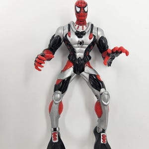 May include: A Spider-Man action figure in a diving suit. The figure is red, black, and silver with a spider emblem on the chest. The figure has black flippers on its feet and is posed with its arms outstretched.