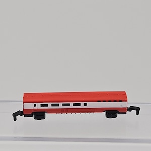 May include: A miniature model passenger train car featuring a bright red roof and lower section with a wide white horizontal stripe running along the middle. The car has a series of small rectangular windows and black plastic wheels and couplers.