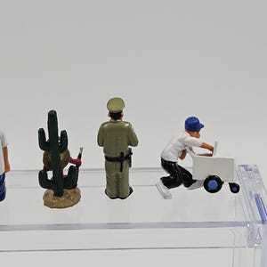 Może przedstawiać: Miniaturowe figurki kaktusa, policjanta i osoby pchającej biały w&oacute;zek. Figurki znajdują się na przezroczystej p&oacute;łce na białym tle. Kaktus jest zielony i brązowy, policjant ma zielony mundur, a osoba białą koszulę i czarne spodnie.