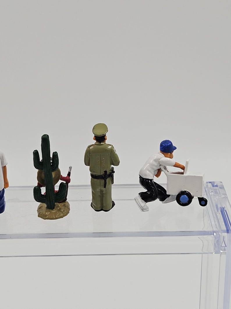 Może przedstawiać: Miniaturowe figurki kaktusa, policjanta i osoby pchającej biały w&oacute;zek. Figurki znajdują się na przezroczystej p&oacute;łce na białym tle. Kaktus jest zielony i brązowy, policjant ma zielony mundur, a osoba białą koszulę i czarne spodnie.