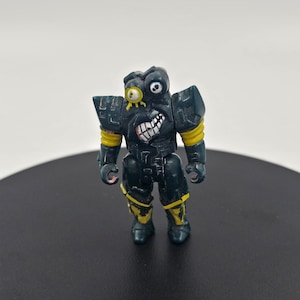 Könnte beinhalten: Eine dunkelgrüne und gelbe Plastik-Actionfigur eines Roboters mit einem großen Auge und einem breiten Grinsen.