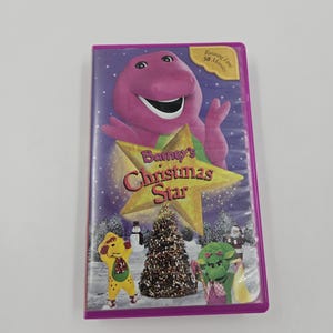 May include: A purple VHS tape featuring Barney the dinosaur with the title "Barney's Christmas Star." The cover shows Barney with a gold star, a Christmas tree, and other characters. The running time is 50 minutes.
