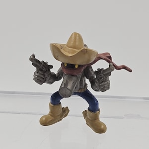 May include: A small figurine of a cowboy character, holding two guns. The figure wears a tan sombrero, a red scarf, a gray jacket, blue pants, and tan boots. The figure is posed as if in action.