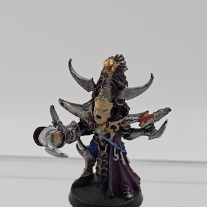 May include: A miniature figurine of a fantasy character with a pale green face, silver horns, and a purple robe. The figure is painted with metallic silver, gold, and red accents, and stands on a black base. The character has multiple arms and weapons.