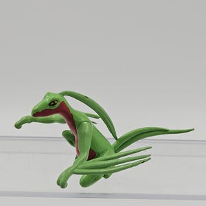 May include: A green and red Sceptile figurine, a Grass-type Pokémon. The figure is in a dynamic pose, with its arms outstretched and its long, leaf-like tail extended. The figure has a red chest and head markings.