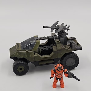 May include: A green and black toy vehicle with large wheels and a mounted gun, accompanied by an orange action figure holding a black weapon. The toy set is designed to resemble a military vehicle.