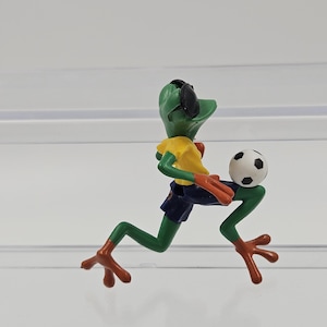 Puede incluir: Una figura de rana verde con una camiseta amarilla, pantalones cortos azules y gafas de sol, corriendo mientras sostiene un balón de fútbol. La rana tiene pies y manos marrones, y el balón es blanco y negro.