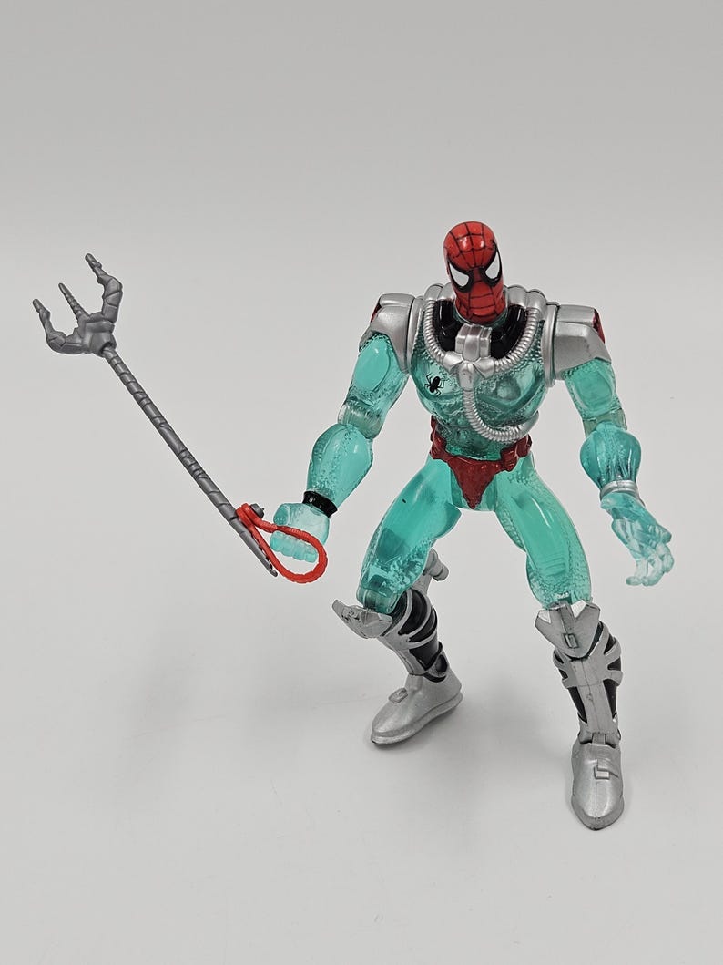 May include: A Spider-Man action figure with a red and black mask, a transparent teal body, and silver armor. The figure is holding a silver trident with a red handle. The figure is in a crouching pose.