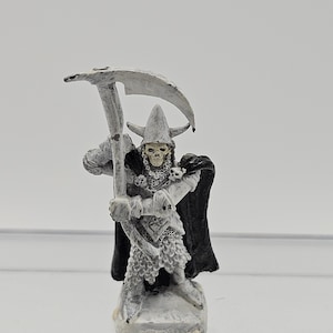 Puede incluir: Figura en miniatura de un guerrero esquelético con armadura plateada, sosteniendo una gran guadaña. La figura lleva un casco con cuernos y una capa negra. La figura está pintada en tonos grises y blancos, con detalles en la armadura y el cráneo.