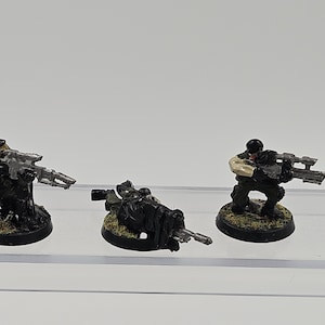 May include: Three miniature soldier figurines mounted on circular black bases. Two figures are in a kneeling position aiming silver rifles, while the center figure is in a prone position aiming a rifle. All figures wear black tactical gear.