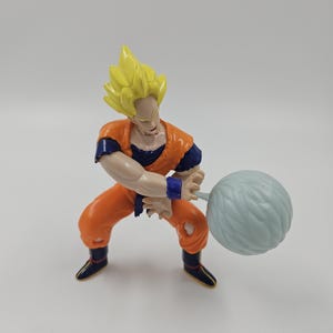 May include: A plastic action figure of a character with spiky yellow hair, wearing an orange and blue outfit, and black boots. The figure is in a fighting stance, holding a light blue ball-shaped object.