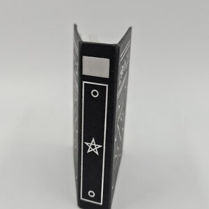 May include: A black book with a silver star and circle design. The spine features a silver rectangle, a circle, a star, and another circle. The book is standing upright against a white background.