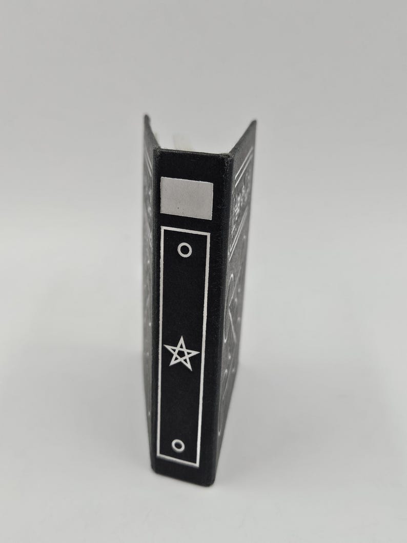 May include: A black book with a silver star and circle design. The spine features a silver rectangle, a circle, a star, and another circle. The book is standing upright against a white background.