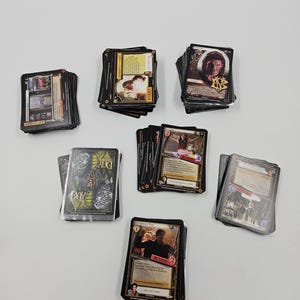 Puede incluir: Varias pilas de cartas coleccionables con bordes negros, cada una mostrando diferentes imágenes y texto. Algunas cartas presentan retratos, otras escenas de acción. Las cartas están dispuestas en un patrón circular sobre una superficie blanca.