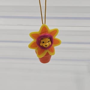 Puede incluir: Un adorno colgante que presenta un personaje de oso amarillo con una expresión de guiño, centrado dentro de un marco con forma de flor de color naranja y amarillo brillante. El personaje está colocado dentro de una pequeña maceta naranja.