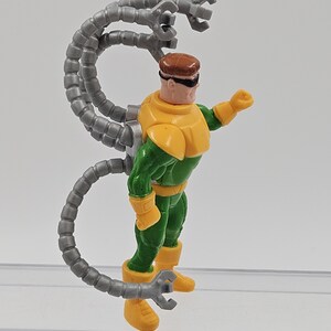 May include: A Dr. Octopus action figure. The figure is wearing a green and yellow suit with a brown helmet and black goggles. Four grey mechanical arms extend from the figure's back. The figure is standing on a clear plastic surface.
