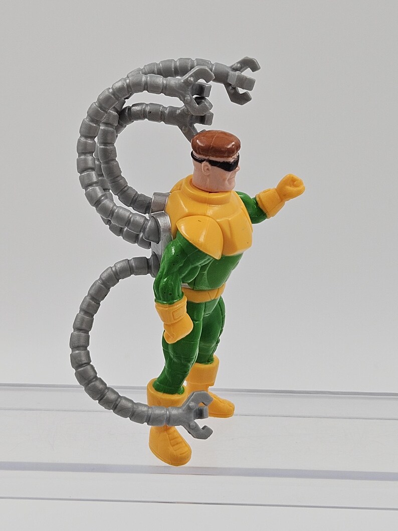 May include: A Dr. Octopus action figure. The figure is wearing a green and yellow suit with a brown helmet and black goggles. Four grey mechanical arms extend from the figure's back. The figure is standing on a clear plastic surface.