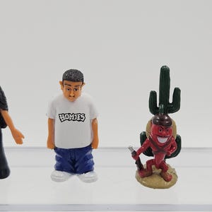 Może przedstawiać: Miniaturowe figurki osoby w białej koszulce z napisem "HOMIES" i niebieskich spodniach oraz czerwonej postaci papryczki chili z pistoletem i kaktusem. Figurki znajdują się na przezroczystej powierzchni.