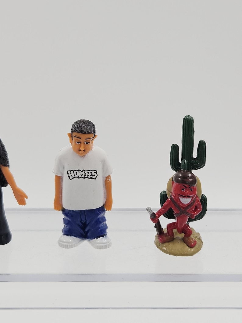 Może przedstawiać: Miniaturowe figurki osoby w białej koszulce z napisem "HOMIES" i niebieskich spodniach oraz czerwonej postaci papryczki chili z pistoletem i kaktusem. Figurki znajdują się na przezroczystej powierzchni.