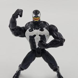 Puede incluir: Figura de acción negra de Venom con un emblema blanco con forma de araña en el pecho. La figura tiene una sonrisa amenazante con dientes amarillos y un símbolo de araña blanco en el pecho. La figura posa con un brazo levantado en un puño.