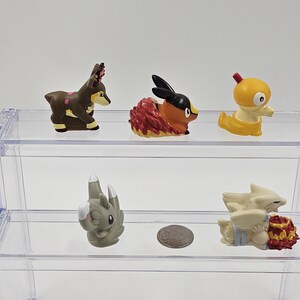 May include: Five small plastic figurines of Pokemon characters displayed on a clear acrylic shelf. The figurines are a brown deer-like Pokemon, a red and orange fire Pokemon, a yellow bird-like Pokemon, a grey rock Pokemon, and a white and red ice Pokemon.