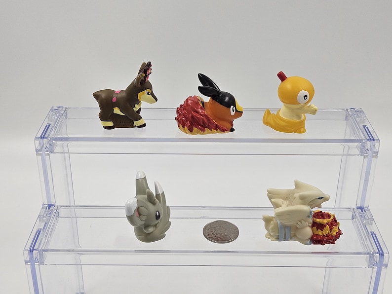 May include: Five small plastic figurines of Pokemon characters displayed on a clear acrylic shelf. The figurines are a brown deer-like Pokemon, a red and orange fire Pokemon, a yellow bird-like Pokemon, a grey rock Pokemon, and a white and red ice Pokemon.