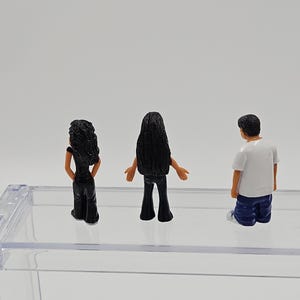 Może przedstawiać: Trzy miniaturowe figurki. Dwie postacie mają długie czarne włosy i noszą czarne stroje. Trzecia postać ma kr&oacute;tkie czarne włosy, biały t-shirt i niebieskie szorty. Figurki stoją na przezroczystej, prostokątnej powierzchni.