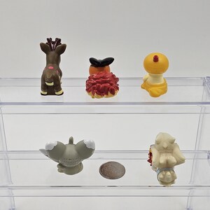 May include: Five small plastic figurines on a clear plastic shelf. The figurines are of various colours and designs, including a brown deer, a red and orange creature, a yellow creature, a grey creature, and a white creature.
