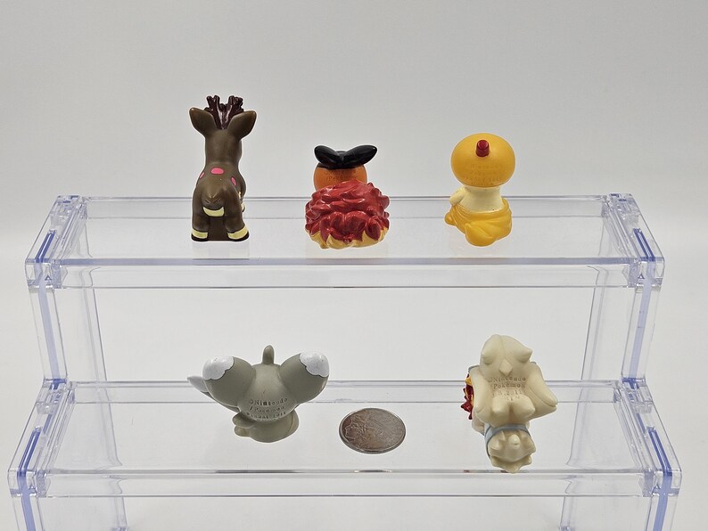 May include: Five small plastic figurines on a clear plastic shelf. The figurines are of various colours and designs, including a brown deer, a red and orange creature, a yellow creature, a grey creature, and a white creature.
