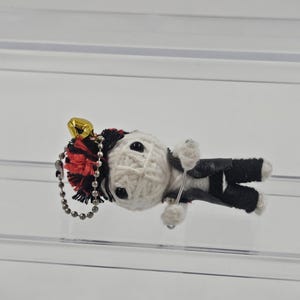 May include: A handmade voodoo doll keychain. The doll is made of white yarn with black button eyes and a black and red tassel. It wears a black vest and pants. A small gold bell hangs from the keychain.