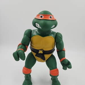 Op de afbeelding: Een groen actiefiguur van een Teenage Mutant Ninja Turtle, met een oranje masker, een gele borstkas en een zwarte riem met een witte "M". Het figuur heeft oranje arm- en beenbanden.