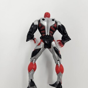 May include: A plastic action figure of a superhero, viewed from the back. The figure has a red head and hands, black arms and legs, and a silver torso. Red and silver accents are visible throughout the design.