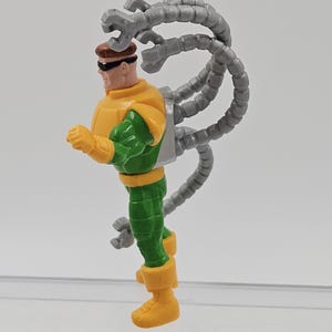 May include: A Dr. Octopus action figure. The figure is wearing a yellow and green outfit with grey mechanical arms extending from its back. The figure has brown hair and sunglasses.